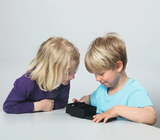 The picture shows two children, a girl and a boy, looking attentively at a black box that is on a table. They seem curious and interested in what is inside the box while working together on the experiment project.