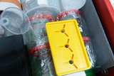 The image shows a collection of experimental materials, including a yellow plastic piece with three openings and various transparent containers. These utensils are arranged in a box-shaped storage unit used for educational purposes.