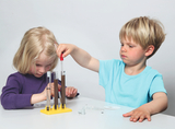 The picture shows two children experimenting with test tubes on a table. A girl with blonde short hair is concentrating on the tubes, while a boy with blonde hair is using a pipette to add liquid to a tube.