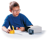The picture shows a boy looking at a white table with a flashlight and a yellow holder. Next to him is a gray experimental device with an image projection that supports him in his experiments.