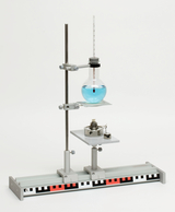 The picture shows a chemical experiment device consisting of a tripod with a glass bottle filled with blue liquid and another container on a platform below. The apparatus is mounted on a stable base frame that allows experiments to be carried out precisely.