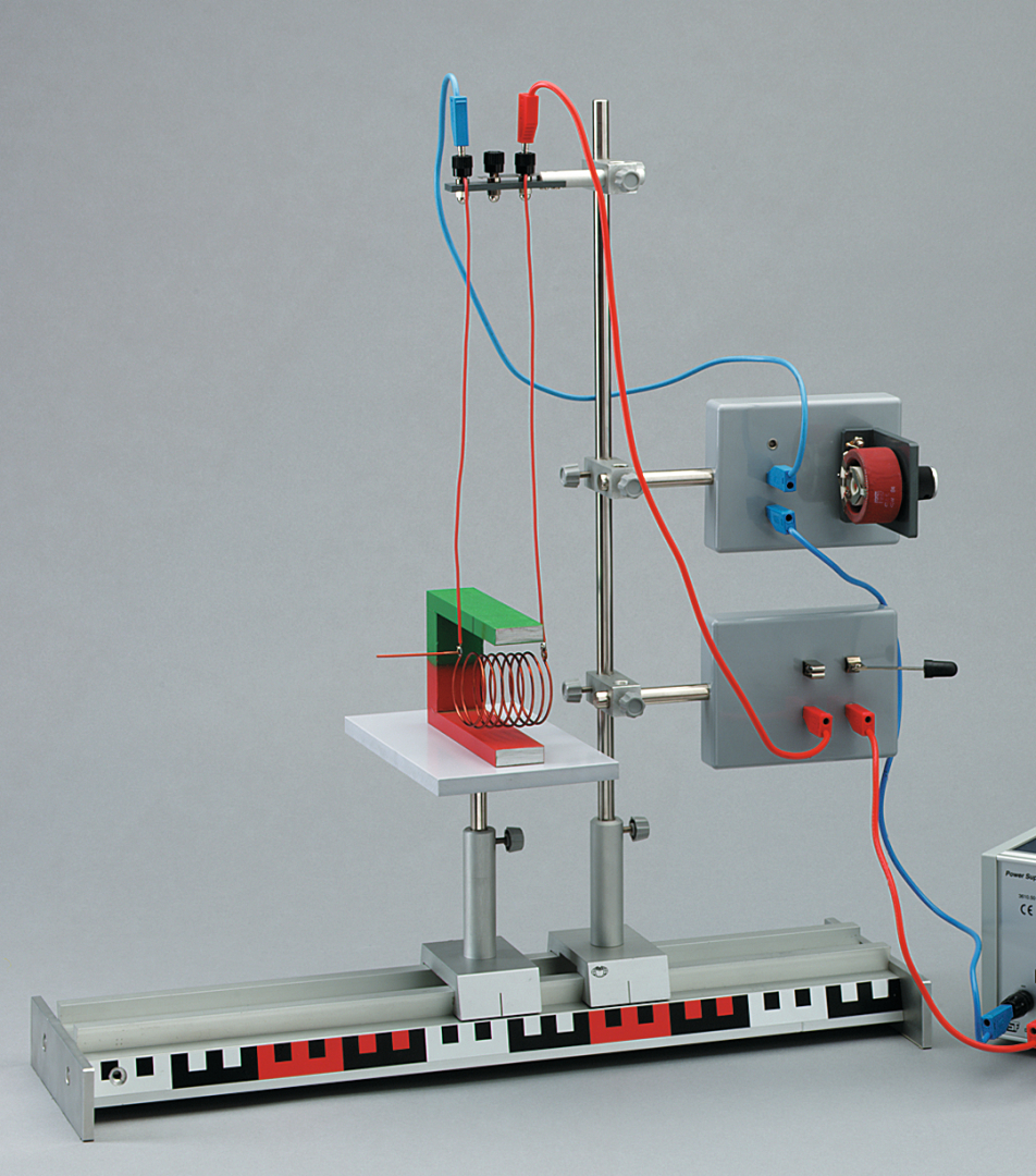 The picture shows an experimental device for demonstrating electromagnetic principles. It consists of an arrangement with coils, a magnet and various connectors for electrical cables mounted on a stable platform.