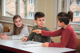 The picture shows three children sitting at a table and communicating with each other. The children seem to be deeply engaged in a conversation while looking at a sheet of paper in front of them.