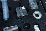The picture shows a collection of experimental materials arranged in a foam organizer. Among other things, you can see a syringe, various parts, a glass container with a chemical substance and short cables.