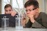 The picture shows two students working in an experimental environment. The boy is focusing on a device, while the girl curiously holds a black cardboard box, presumably to cover part of the experiment.