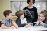 The picture shows four children in a classroom working on an experiment. One girl is reading from a book while another child is working with an apparatus to observe a chemical reaction.