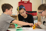 The picture shows three children working together on craft projects. They are using colored paper and seem to be engaged and focused on the task.