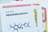 The picture shows instructions for molecule construction kits from Cornelsen Experimenta, which are used to design and investigate molecular structures. Additional materials and information about the experiments are visible in the background.