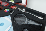 The picture shows a black foam tray with various experiment materials, including a magnifying glass and a spoon. Also visible are colored elements such as a color wheel and some small objects that can be used for experiments.