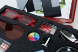 The picture shows an experiment kit in a red case with a foam insert. It contains various items such as red-tinted glasses, a color game and other tools that can be used for experimental purposes in education.