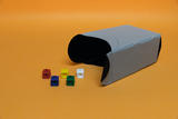 The picture shows a gray, curved box with a black interior lying on an orange background. Next to it are several colored, square building blocks in red, yellow, green, blue and white.