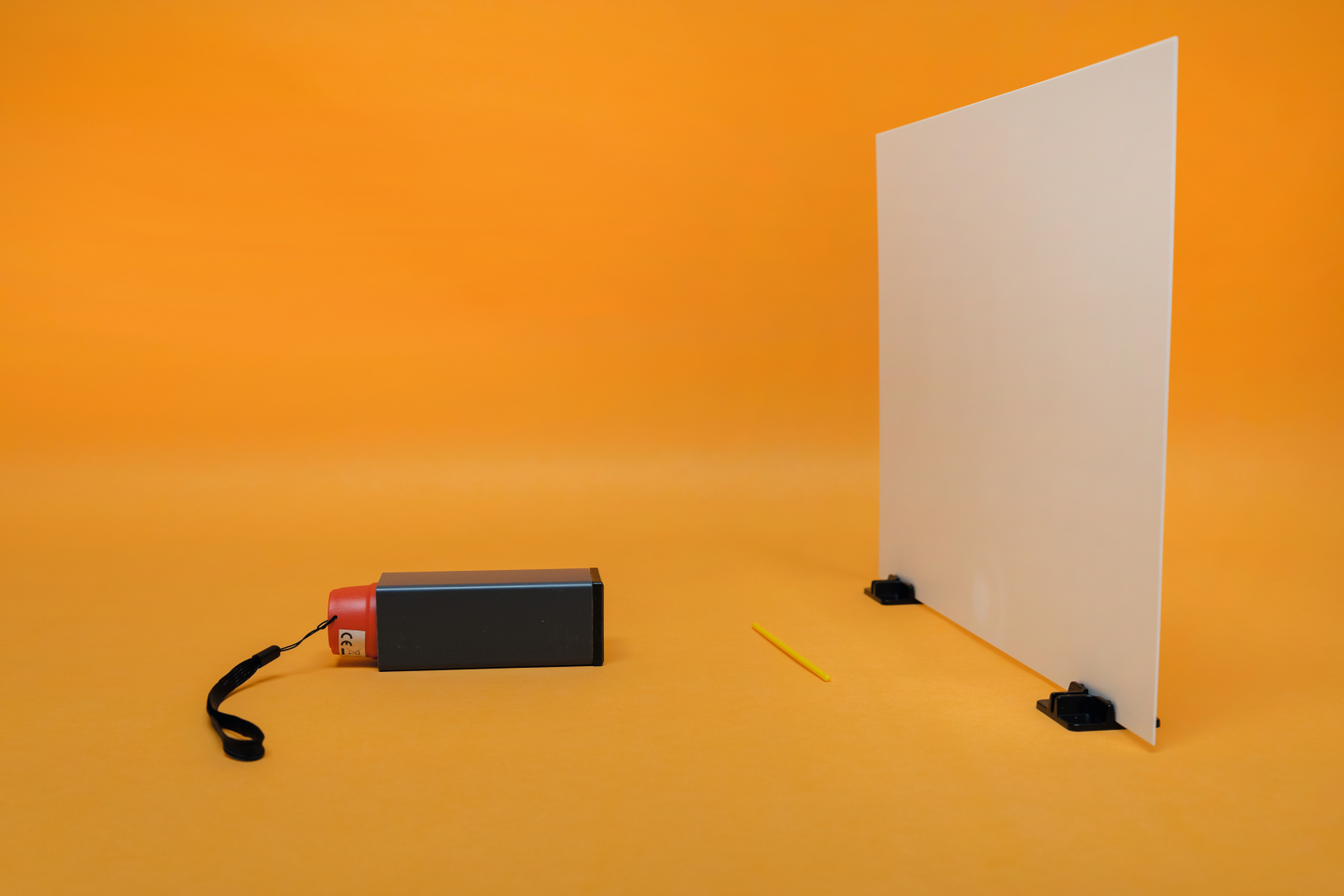 The picture shows an experiment block consisting of a black device with a red head and a white viewing screen supported by black brackets. Next to the equipment lies a yellow pen on the orange background.