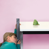 The image shows a child leaning sideways on a table, looking intently at a green stuffed animal sitting on the tabletop. The background is a soft pink, creating a friendly and inviting atmosphere.