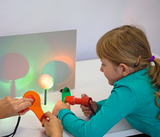 The picture shows a child experimenting with a red flashlight-like device and an orange lens, while a second person works with an orange lens. On the table are two light sources that project different colors of light.