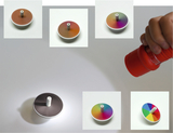 The picture shows various spinning tops with different color patterns arranged on a table. A hand holds a flashlight pointed at one of the tops to illustrate the play of colors.