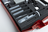 The image shows a red suitcase filled with different shaped and dimensional experimental materials arranged in gray foam inserts. Among other things, you can see transparent containers, plates and a tool that can be used for scientific experiments in educational settings.