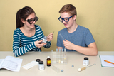 The picture shows two young people working with experimental materials at a table. They are using test tubes and various chemicals while actively exchanging ideas about how to conduct experiments.