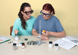 The picture shows two young women wearing safety glasses working at a table with experimental materials. On the table surface you can see various chemicals, a burner and a textbook for experiments.
