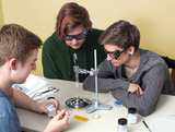 The picture shows three students working concentratedly at an experiment table. They use laboratory equipment, including a heat source, test objects in containers, and safety goggles, to conduct chemical or physical experiments.