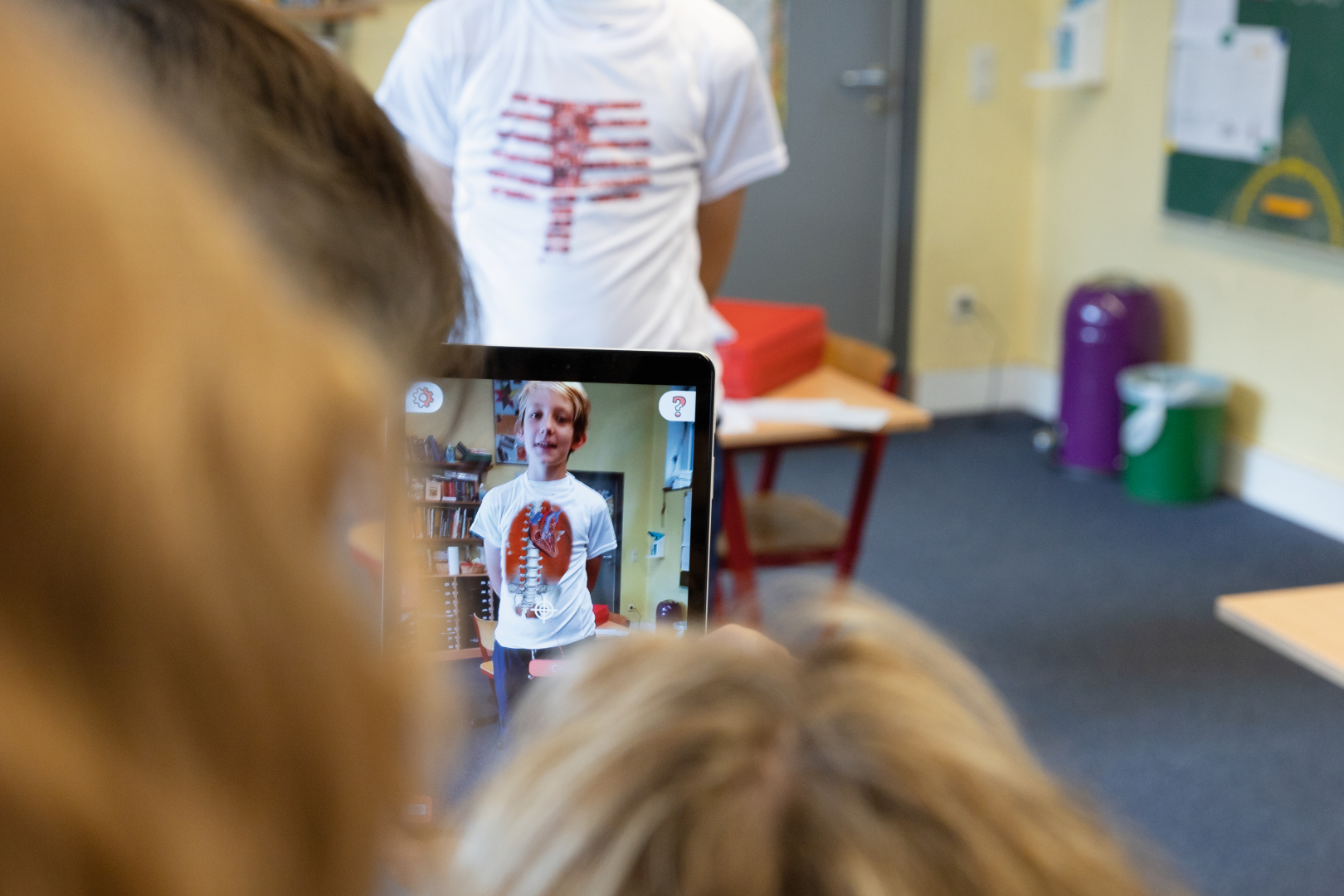 The picture shows a child wearing a t-shirt with an anatomical motif and looking at a tablet. In the background there are other students and a classroom with shelves and a green chalkboard area is visible.