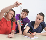 The picture shows three students working on an experiment. One student is holding a syringe over a device with rollers, while the other two are watching carefully and taking notes.