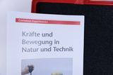 The picture shows a booklet entitled “Forces and Movement in Nature and Technology” by Cornelsen Experimenta. It is lying on a black, foam-like base and shows a hand with a stopwatch device.