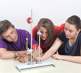 The picture shows three young people working together on an experimental setup with a lever system. They watch carefully as a weight is placed on a device with rollers and a hook.