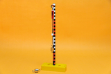 The image shows an experimental device with a vertical rod holding a spring balance and a heavy block. The base is bright yellow and provides stability for the experiment, which probably illustrates physical principles.