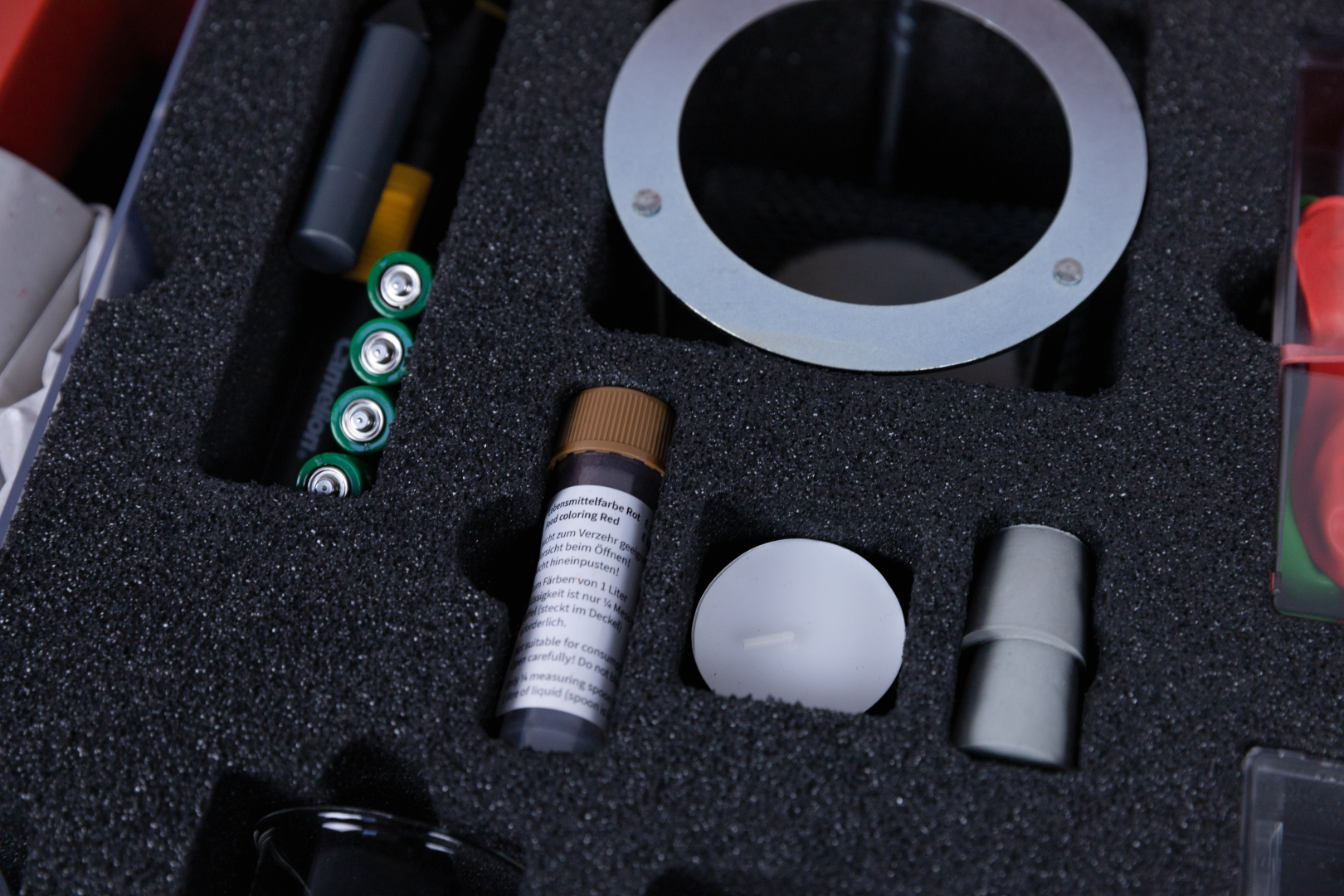 The picture shows a set of experiment materials arranged in a foam tray. The items included include batteries, a colorant, a tea light and various accessories for educational experiments.