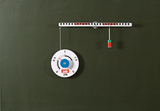The image shows a mechanical weighing measurement with a scale that indicates the weight of a suspended object. A colored cylinder hangs on a string and acts as a weight while the indicator calibrates to 2 Newtons.