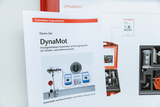 The picture shows an information sheet about the DynaMot Demo Set, a hand-operated generator ideal for experiments in school lessons. In the background, the associated equipment is shown in a red case, which is used for student and teacher experiments.