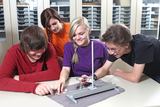 The picture shows four young people conducting an experiment at a table. They are gathered around a scientific device and are observing intently while one of them places a small object on it.