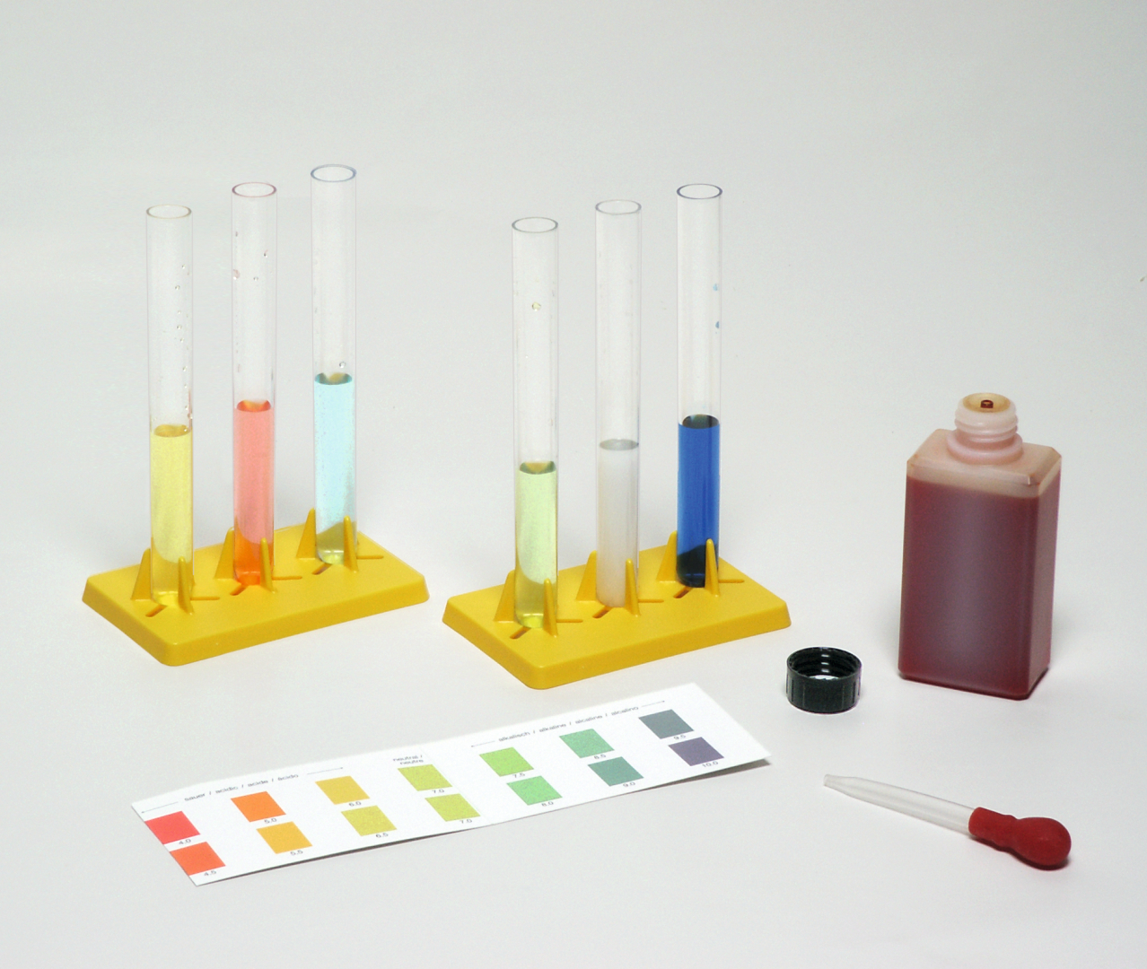 The picture shows several test tubes in a yellow stand, each filled with different liquids. Next to them is a pipette and a bottle with red liquid, as well as a pH test strip with colored scales.