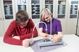The picture shows two teenagers working together on an experiment. They are using a chemical apparatus set, with the boy holding a liquid in a beaker and the girl working at the experiment table.