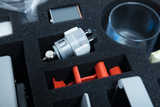 The picture shows a compartment layout in a foam case in which various experimental materials are housed. The visible objects include a silver hydraulic cylinder, red plastic parts and a transparent glass.