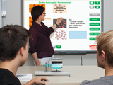 The picture shows a teacher explaining the topic of direct use of solar energy on an interactive screen. Two students are watching the lecture and working on an experiment dealing with temperature and absorption.