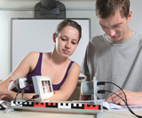 The picture shows two students working on an experimental setup. They are using various equipment to carry out an experiment, with the female student focusing on the setup and the male student studying instructions.