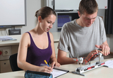 The picture shows two students working on an experimental setup that appears to be related to wind power. The female student is making notes while the male student is examining the electrical components of the model.
