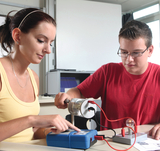 The picture shows two students conducting an experiment with an electrical circuit. The female student is operating a meter while the male student is connecting to a light bulb to explore how electricity works.