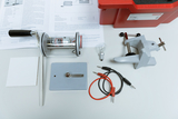 The image shows a selection of experiment materials including a motor, light bulb, wires and a clamp on a table. Additionally, an information sheet and storage box are visible, summarising the materials for educational purposes.