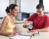 The image shows two students in a classroom working on an experiment. The boy is operating a device while the girl is taking notes and smiling, indicating an engaged learning atmosphere.