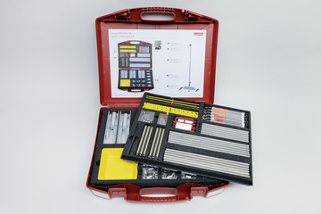 The picture shows a red suitcase equipped with various experimental materials for teaching. The inside contains an overview of the parts included as well as an orderly arrangement of rods, brackets and accessories for physics experiments.