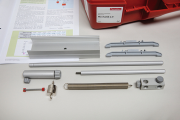 The image shows various components of a mechanics kit, including metal and plastic parts and an instruction manual with diagrams and formulas. These components are designed to help students understand physics concepts.