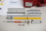 The picture shows an experiment kit with various components, including aluminum profiles, a force gauge, and connecting elements that are used for physical experiments on mechanics. In addition, instructions are included that explain how to use the materials.