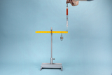 The picture shows an experimental device for carrying out physical measurements. A scale with a hook weight hangs from a yellow beam mounted on a stable base, while a handle for adjustment is visible.