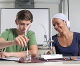 The picture shows a student and a teacher working together on an experiment. The student is focusing on pipetting a liquid into a beaker while the teacher watches him, smiling.
