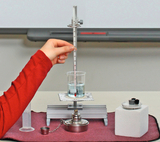 The picture shows an experimental setup with a beaker, a thermometer and a heater on a table. A hand holds the thermometer in a beaker of water while a gray pad and other laboratory equipment are placed nearby.