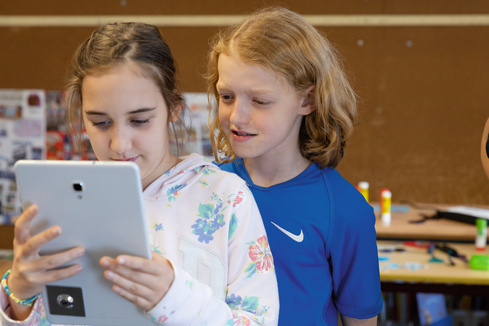 The picture shows two children looking at a tablet together. The atmosphere seems curious and friendly, with artistic materials and posters visible in the background, suggesting a creative learning environment.