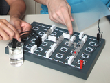 The image shows an experiment board with various electrical components and a liquid source. Two people are manipulating the setup to make electrical connections and possibly conduct an experiment.