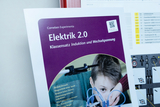 The picture shows a teaching material called &quot;Electricity 2.0&quot;, which deals with the topics of induction and alternating current. It also shows a boy working with experimental equipment to research electrical phenomena.